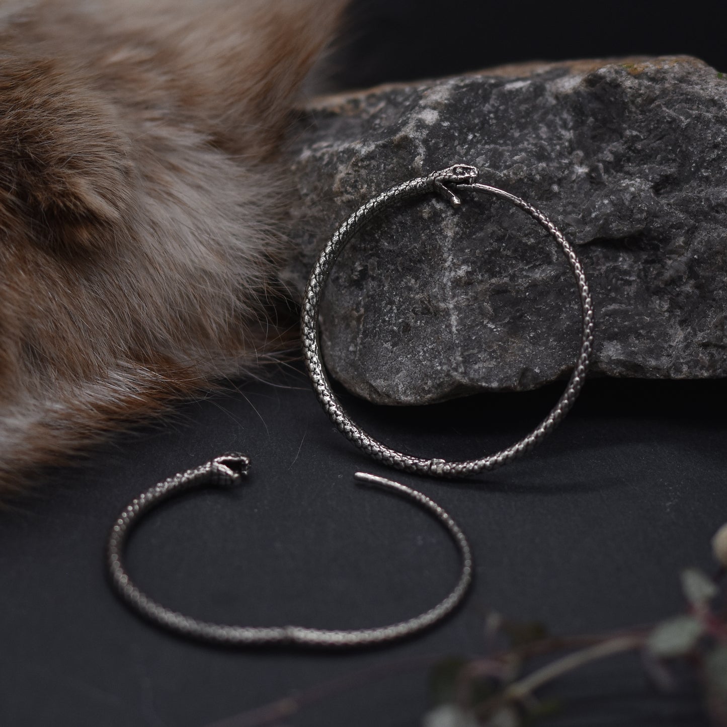 A pair of silver-colored brass hoop earrings with a textured design, displayed on a dark surface with a rock and a piece of fur in the background.