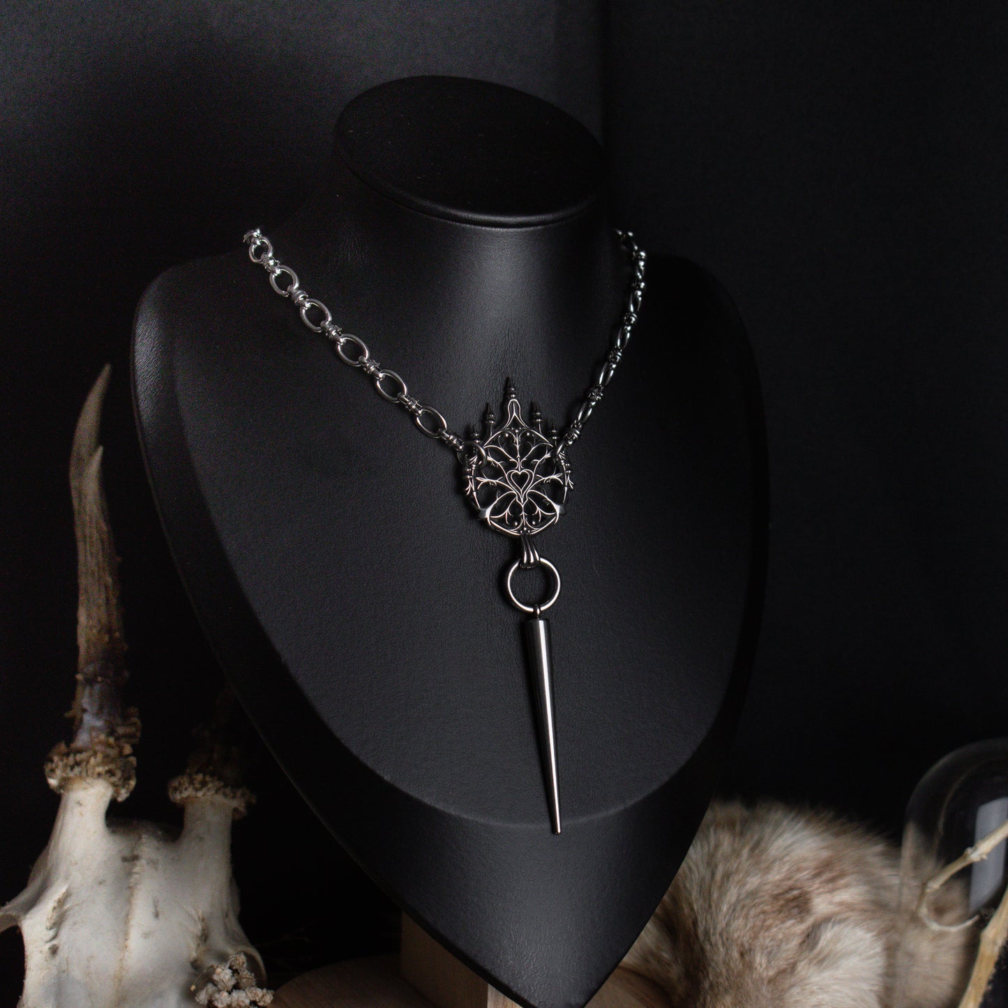 A silver gothic style pendant chain necklace displayed on a black mannequin bust, featuring a detailed ornament
and a long pointed spike, with decorative animal skull and fur props against a dark background.