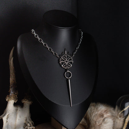A silver gothic style pendant chain necklace displayed on a black mannequin bust, featuring a detailed ornament
and a long pointed spike, with decorative animal skull and fur props against a dark background.