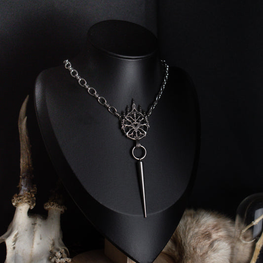 A silver gothic style pendant chain necklace displayed on a black mannequin bust, featuring a detailed ornament
and a long pointed spike, with decorative animal skull and fur props against a dark background.