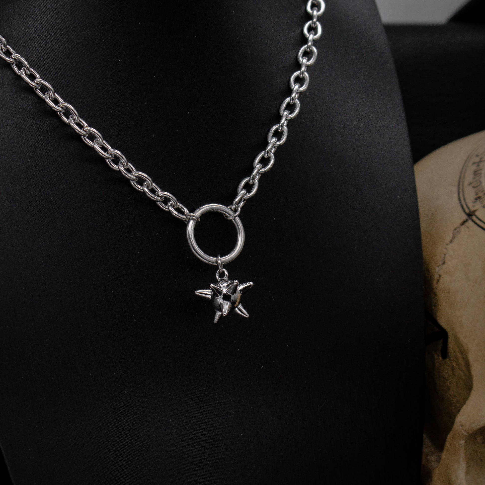 A silver medieval flail necklace with a chunky chain and a circular pendant, displayed on a black stand with a skull in the background.